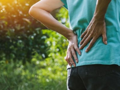 A person clutches their lower back as if in pain. They are outside with greenery in the background.